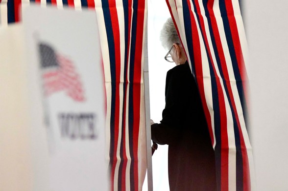 A voter casts a ballot at a polling place, partially obscured by red, white, and blue striped curtains.